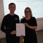 A prize-winning student stands with Emma Fielding, they are holding the student's certificate and smiling.