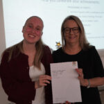 A prize-winning student stands with Emma Fielding, they are holding the student's certificate and smiling.