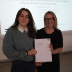 A prize-winning student stands with Emma Fielding, they are holding the student's certificate and smiling.