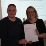 A prize-winning student stands with Emma Fielding, they are holding the student's certificate and smiling.