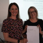 A prize-winning student's parent stands with Emma Fielding, they are holding the student's certificate and smiling.