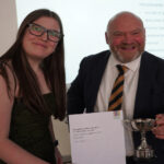 A prize-winning student stands with Bill Revans, they are holding the student's certificate and trophy, and smiling.