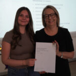 A prize-winning student stands with Emma Fielding, they are holding the student's certificate and smiling.