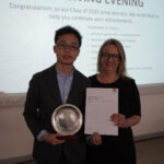 A prize-winning student stands with Emma Fielding, they are holding the student's certificate and certificate, and smiling.