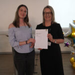 A prize-winning student stands with Emma Fielding, they are holding the student's certificate and smiling.