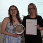 A prize-winning student stands with Emma Fielding, they are holding the student's certificate and certificate, and smiling.