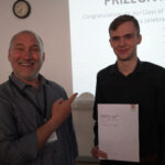 A prize-winning student is holding his certificate and stands with a staff member, who is pointing at smiling at him.