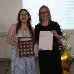 A prize-winning student stands with Emma Fielding, they are holding the student's certificate and trophy, and smiling.