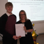 A prize-winning student stands with Emma Fielding, they are holding the student's certificate and smiling.