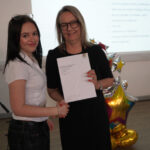 A prize-winning student stands with Emma Fielding, they are holding the student's certificate and smiling.