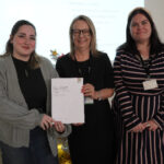 A prize-winning student stands with Emma Fielding and a staff member, they are holding the student's certificate and smiling.