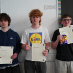 Three students stand holding certificates in front of a classroom display.