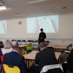 A student presents a mobile app slide to judges seated at classroom tables.