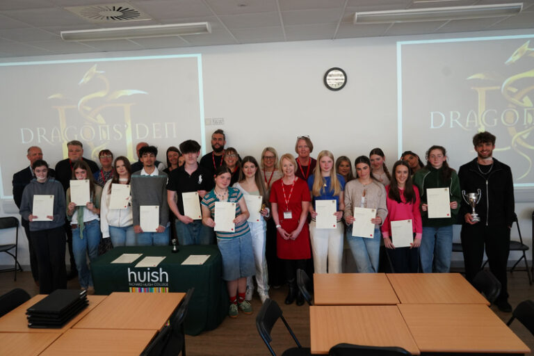 A large group photo of students and business representatives holding certificates in front of “Dragons’ Den” screens.