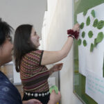 Two individuals engage in an activity, attaching paper leaves with written pledges to a tree illustration on a bulletin board. The scene is indoors.