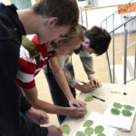 A group of people standing around a table are engaged in writing pledges on green paper leaves. The setting appears to be indoors, near a staircase