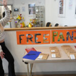 A person is doing jazz hands and standing next to a display table with a colourful sign reading "FREE PANTS." The table has paper packets of pants, leaflets and a clipboard. In the background, there are balloons and informational posters on the walls.
