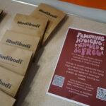 The image shows a display of Modibodi products in brown packaging on a tabletop. Next to them is a sign advocating for free feminine hygiene products, highlighting cost savings and environmental benefits. Two QR codes are at the bottom of the sign.
