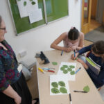 A person with stands next to a table where two individuals are drawing on green leaves. They are writing pledges on them.