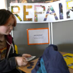 A person is sitting at a table working with fabric pieces. Behind them is a sign that says "REPAIR" and another with the name "azengear." There’s clothing on the table and part of a coat.