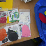 The image shows several packets of Repair Patches in different colours and shapes on a wooden table. The packets include patches in red, black, white, and pink. A blue fabric with a red and yellow patch partially attached is also visible.