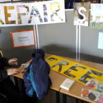 A person sits at a table in a repair shop area. Above the table, there are large signs reading "REPAIR SHOP" and there is also a sign on the table that reads "FREE." The table has various items and a jacket on it. Information about Azengear is displayed on the boards behind.