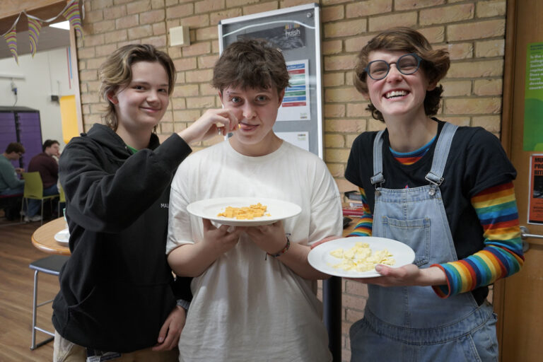 Three people smiling indoors, with two holding plates of vegan cheese. A brick wall and informational poster are in the background.