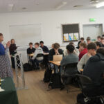 A speaker addresses seated students in a classroom while trophies sit on a table nearby.