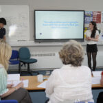 Two students present a product concept on a screen to an audience in a classroom.