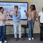 Four students stand at the front of a classroom presenting a project titled “MindHive Solo pods.”