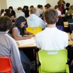 A busy classroom with students seated at tables. One is writing on paper.