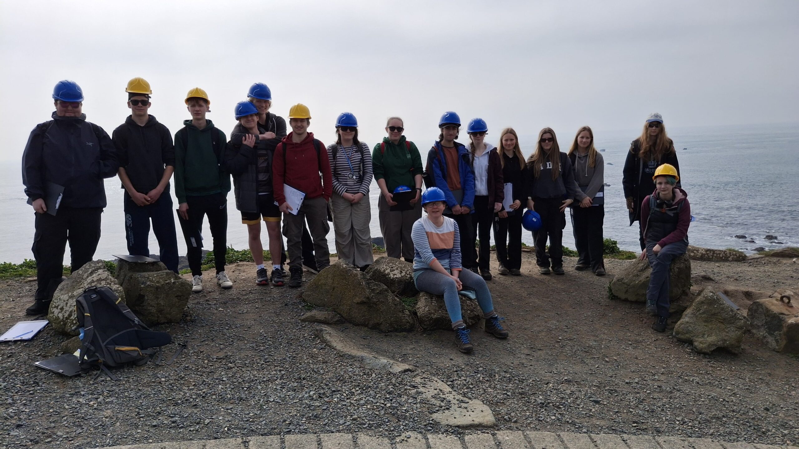 Huish Geology students and staff with hard hats on, stood in front of the coast. There are some large rocks in front of them.