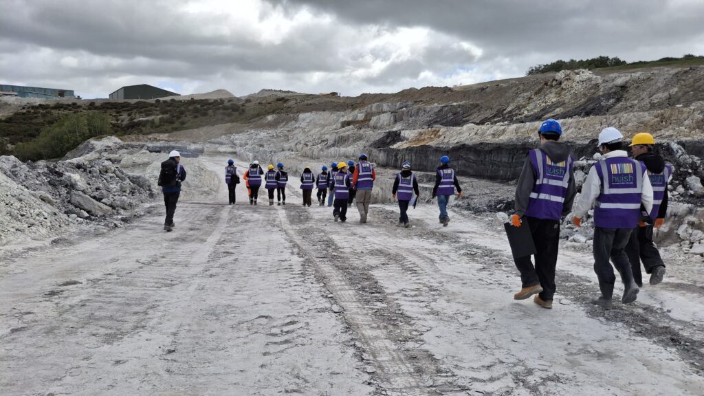 A scene of a quarry with lots of students walking down hill. They are wearing hard hats and purple Huish branded hi-vis jackets.