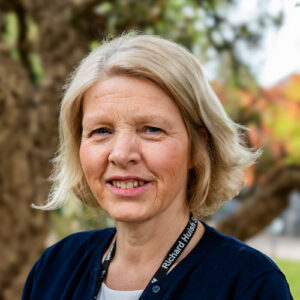 The head and shoulders of a woman with short blonde hair. She is wearing a white t-shirt, blue cardigan, and a black lanyard.