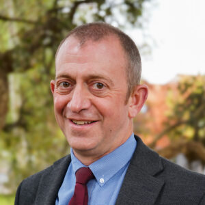 The head and shoulders of a man with short hair. He is wearing a blue shirt, burgundy tie and grey suit.
