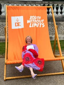 A Huish staff member sitting on a large orange deck chair with DofE branding. She is wearing a bright dress with poppies on.