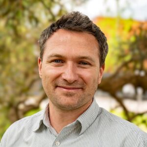 The head and shoulders of a man with short hair. He is wearing a white pin striped shirt.