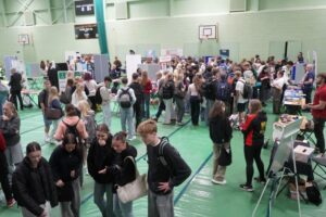Lots of people milling about in a sports hall, at a fair. There are multiple stands that students are gathered round.