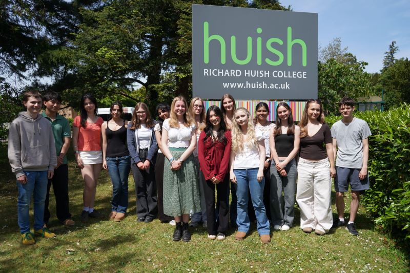 Multiple students stand smiling in front of a large Huish sign. They are in a grassy area with trees behind. It is sunny.