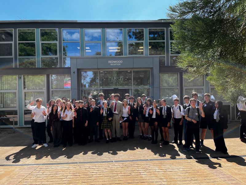 A large group of students stood close together in front of the Huish reception. Most wear school uniform.