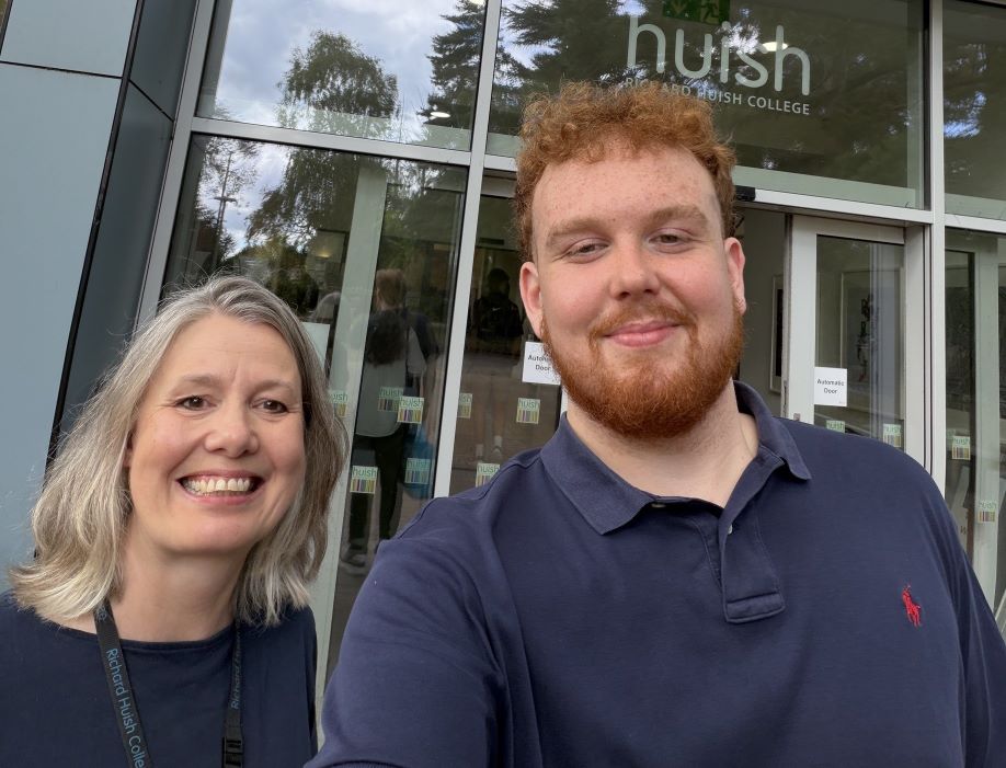 A teacher and former student are standing smiling in front of the entrance to a building labelled "Redwood Reception" at Richard Huish College. Trees are reflected in the glass behind them.