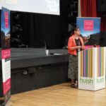 A business or university representative giving a talk in a theatre. She stands behind a podium.