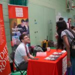 A university or business representative chatting to students at an exhibition stand.