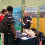 A university representative chatting to students at an exhibition stand.