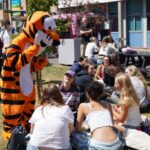 A busy grassy area outside with lots of students milling about chatting. The Huish tiger is also there.