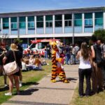 A busy grassy area outside with lots of students milling about chatting. The Huish tiger is also there.