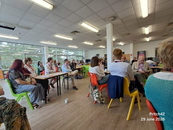 People are sat at tables on colourful chairs. They are in a big space that looks like a classroom.