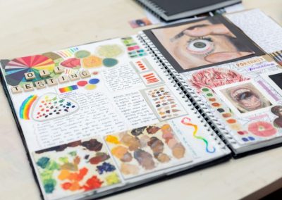 A close up of a student's art work book. There is a title made from scrabble pieces, saying 'Oil Testing'. There is writing and splodges of paint on one page, and images, including of an eye and mouth, on the other page.