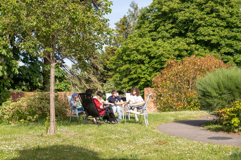 Students enjoying Oak House’s outdoor garden space, relaxing around a table in a peaceful setting with mature trees, a lawn and pathway