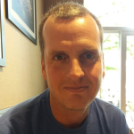 A man with short hair and a blue top smiles with his mouth closed.